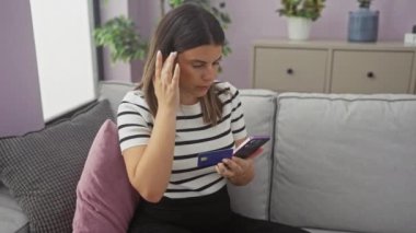 Genç esmer kadın oturma odasında kart ve telefon tutuyor, muhtemelen internetten alışveriş yapıyor..