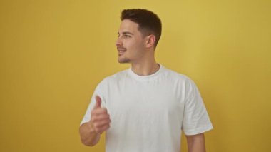 Beyaz tişörtlü neşeli, genç İspanyol adam, neşeyle işaret ediyor ve sarı izole edilmiş arka plana bakıyor.