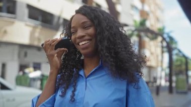 Kıvırcık saçlı genç Afro-Amerikalı kadın şehir sokaklarında gülümseyen, açık havada mavi bir gömlek giyen telefonundaki sesli mesajı dinliyor..
