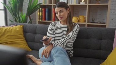 İspanyol kadın modern bir oturma odasında uzaktan kumandayla kanepede uzanıyor.