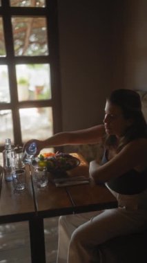 Düşünceli genç bir kadın, modern bir restoran ortamında masaya sağlıklı bir salata menüsü koyuyor..  