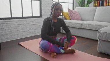 Örgü ören Afrikalı Amerikalı kadın modern bir oturma odasında pembe bir paspasın üzerinde yoga yapıyor..