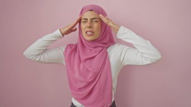 Tesettürlü genç İspanyol kadın, elleri başının üstünde, migrenle savaşıyor. Acı, izole pembe arka planda yankılanıyor. Stresli bir ağrının bedeli vardır..