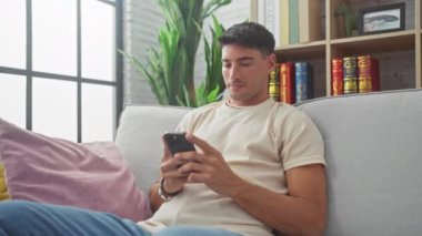 İspanyol asıllı mutlu bir adam oturma odasındaki koltukta akıllı telefon kullanıyor, modern yaşam tarzı ve bağlılık sergiliyor.