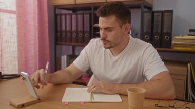 Genç İspanyol bir adam, arka planda dosya klasörleri olan modern bir ofiste akıllı bir telefona bakarken deftere notlar yazıyor..