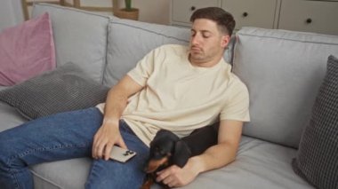 Genç İspanyol bir adam modern bir oturma odasında tekel köpeğiyle dinleniyor, rahat gri bir kanepede uzanırken akıllı telefon kullanıyor..