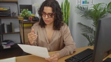 Ofisteki genç İspanyol kadın ofis ekipmanları ve yeşilliklerle çevrili bir belgeyi okumak için gözlüklerini çıkarıyor..