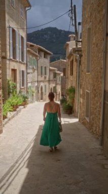 Yeşil elbiseli kadın Valldemossa, Mallorca, İspanya 'da dar bir taş sokakta yürüyor. Arka planda saksı bitkileri ve dağları olan büyüleyici eski binalarla çevrili.