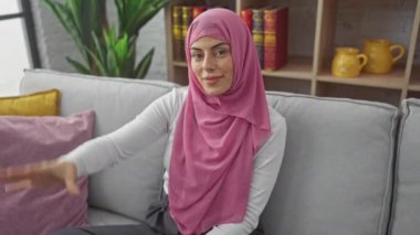 Neşeli genç İspanyol kadın, evinde koltukta tesettür içinde, kendine güvenen bir gülümseme ile ışıldıyor, parmaklarıyla komik bir 