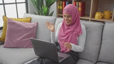 Başörtüsü takan gülümseyen bir kadın rahat bir oturma odasında dizüstü bilgisayarla sohbet ediyor.