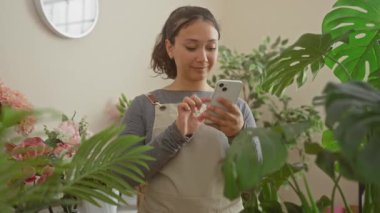 Heyecanlı genç İspanyol çiçekçi kadın çiçekçisinden bir zafer kazandı, başarısını neşeli bir gülümseme ve muzaffer bir ifadeyle kutluyor, akıllı telefonuyla yakalandı..