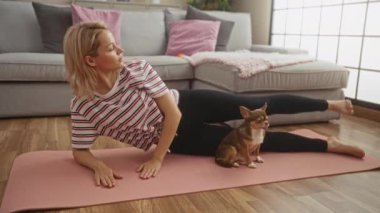 Sarışın genç bir kadın oturma odasında yoga minderiyle egzersiz yapıyor yanında da Chihuahua 'sı var..
