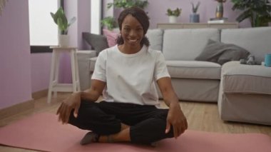 Oturma odasındaki yoga minderinde oturan, rahat bir evin içindeki kamerayla konuşan kıvırcık saçlı Afro-Amerikalı bir kadın..