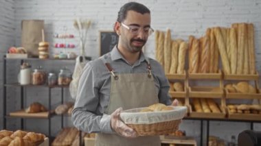 Önlüklü genç bir adam arka planda çeşitli hamur işleri ve baget sergilenen fırında taze ekmek sepeti tutuyor.