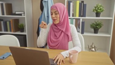 Neşeli, tesettürlü genç İspanyol kadın, neşe içinde gülümsüyor, bilgisayar ekranını işaret ediyor, kendine güveni tam bir jest yapıyor.