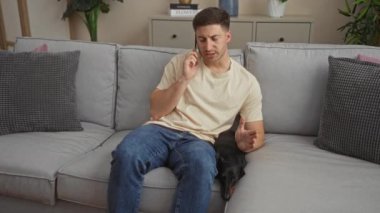 Genç adam kanepede köpeğiyle oturuyor, rahat bir oturma odasında telefonla konuşuyor..