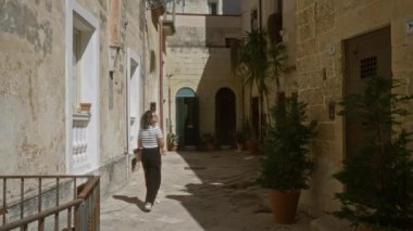 Genç bir kadın Lecce, Puglia, İtalya 'da büyüleyici, dar, güneşli bir sokakta yürüyor. Tarihi taş binalara ve saksı bitkilerine hayran kalıyor..