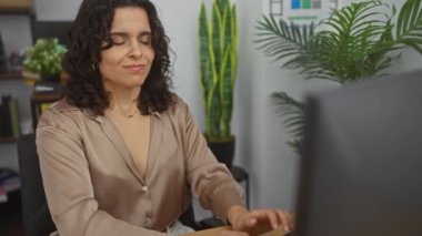 Ofis ortamındaki genç, çekici esmer kadın, arka planda bitki ve raflarla dolu bir masada otururken rahatsız ve boynuna dokunuyor..