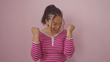 Neşeli genç İspanyol kadın, çizgili tişört giyiyor, pembe arka planda dikiliyor, kendinden emin zaferi heyecanla kutluyor..