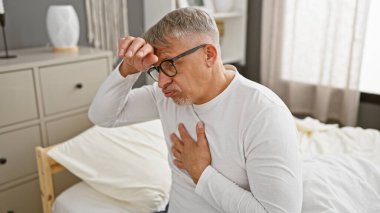 Rahatsızlık içindeki orta yaşlı bir adam iyi ışıklandırılmış bir yatak odasında bir yatağa oturur. Hastalık ya da baş ağrısı önerir..