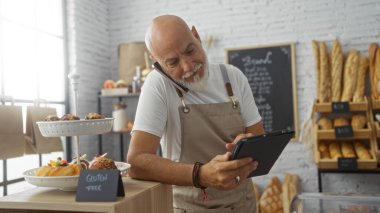 Gri saçlı ve sakallı olgun bir adam, fırında çalışıyor, tableti kontrol ediyor, kapalı bir dükkânda telefonla konuşuyor ve etrafta ekmek ve pastalar sergileniyor..