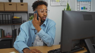 Ofiste çalışan Afro-Amerikalı genç bir adam, arka planda ofis dekoru ve rafları olan bir bilgisayar ekranına bakarken telefonla konuşuyor..