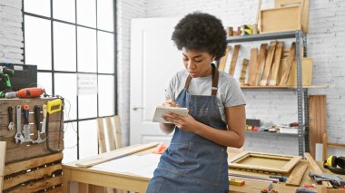 Kot önlüklü odaklanmış siyah bir kadın parlak bir marangozluk atölyesinde notlar alıyor..