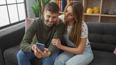 Bir erkek ve bir kadın, parlak bir oturma odasındaki gri bir kanepede akıllı bir telefonla samimi bir anı paylaşıyor..