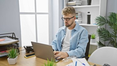 Gözlüklü ve sakallı genç adam modern bir ofiste dizüstü bilgisayarla çalışıyor..