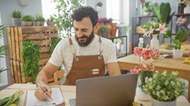 Sakallı adam dizüstü bilgisayarlı ve yeşil bitkilerle çiçekçide yazıyor, olgun, profesyonel bir atmosfer yayıyor..