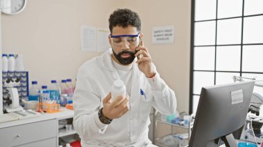 Genç bir adam laboratuarda bir şişeyi incelerken telefonda konuşuyor, güvenlik gözlüğü ve laboratuvar önlüğü takıyor..