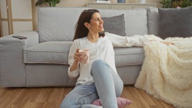 Genç bir İspanyol kadın, rahat bir apartman dairesinde, bir kanepenin yanında oturuyor, elinde bir fincan, ve evde rahat bir anın tadını çıkarıyor..