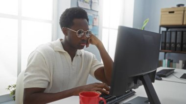 Genç Afro-Amerikalı bir adam bilgisayarın ofisinde çalışıyor. Masasında kırmızı bardakla, gözlüklü ve beyaz gömlekli..