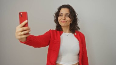 Kırmızı kazaklı ve beyaz bluzlu genç bir İspanyol kadın özgüvenli ve çekici bir pozla beyaz bir arkaplanda selfie çekiyor..