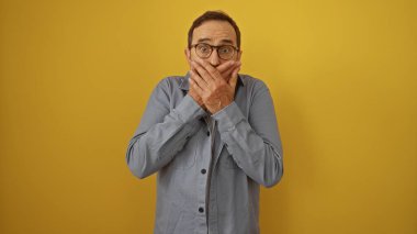 Orta yaşlı bir adam sarı bir arkaplan karşısında izole edilmiş ağzını iki eliyle kapatıyor sürpriz veya şok içinde, gözlük takıyor ve gri gömlek giyiyor..