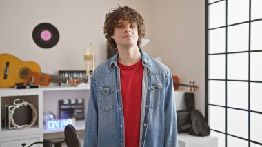 Modern bir müzik stüdyosunda poz veren kıvırcık saçlı yakışıklı genç adam, sıradan bir tarz ve yaratıcı bir ortam sergiliyor..