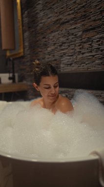 Huzurlu genç bir kadın modern bir banyoda köpük banyosunun keyfini çıkarıyor, taş duvarlar huzurlu bir ortam sağlıyor..