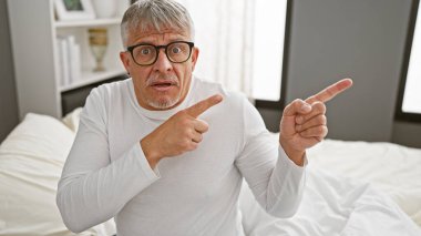 Şaşırmış orta yaşlı gözlüklü bir adam modern bir yatak odası ortamında parmaklarını içeriyi işaret ederek şok ya da keşif yapıyor..