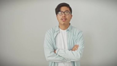 Gözlüklü huysuz genç Çinli adam, şüpheli ve üzgün, kuşkucu bir ifadeyle hoşnutsuzluk gösteriyor ve kollarını kavuşturuyor, beyaz duvarda izole edilmiş..