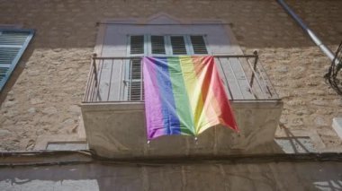 Güneşli bir taş binanın balkonundan sarkan gökkuşağı bayrağı lgbtq gururunu ve dışardaki kapsayıcılığı temsil ediyor..