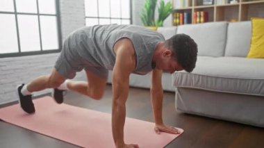 Formda bir genç adam modern bir oturma odasında pembe bir yoga minderi üzerinde egzersiz yapıyor ve evdeki sağlıklı yaşam tarzını gösteriyor..