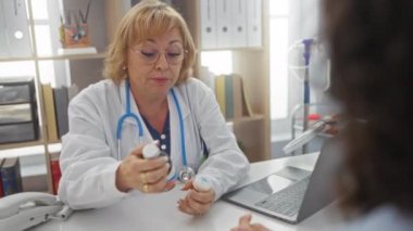 Orta yaşlı kadın doktor, klinik ortamdaki bir hastaya ilaçlarını açıklıyor. Etrafı ofis malzemeleriyle çevrili, iyi organize edilmiş bir sağlık ortamında profesyonel bir etkileşim sergiliyor..