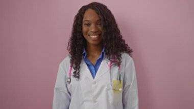 Doktor, Afro-Amerikan üniformalı genç bir kadın, mutlu bir gülümsemeyle duruyor, kendinden emin ve pozitif, pembe arka plana bakıyor, güzel dişlerini gösteriyor.