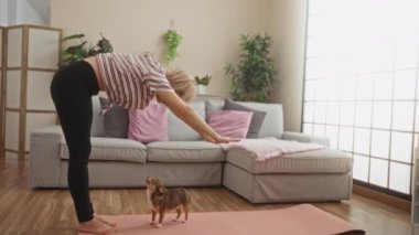 Genç sarışın bir kadın, kapalı alanda yoga yapıyor. Chihuahua 'sı ile, bitki ve doğal ışıkla dolu, rahat bir oturma odasında, pembe bir paspasın üzerinde..