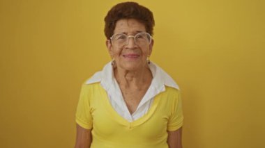 Kısa saçlı, gözlüklü, sarı gömlekli, izole edilmiş sarı arka planda duran, kendine güvenen, yaşlı İspanyol kadın..
