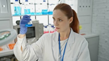 A focused young woman scientist examines a blue liquid in a flask in a well-equipped laboratory. clipart