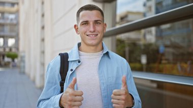 Sırt çantalı, gülümseyen genç İspanyol bir adam şehir koleji ortamında başparmağını kaldırıyor.