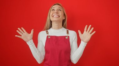 Kırmızı arka planda izole edilmiş beyaz gömleğin üzerine kırmızı tulum giyen, sarı saçlı, gülümseyen beyaz bir kadın..