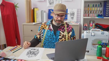 Gözlüklü, sakallı, şık, orta yaşlı bir adam renkli bir terzi stüdyosunda dizüstü bilgisayardan tasarımları inceliyor..