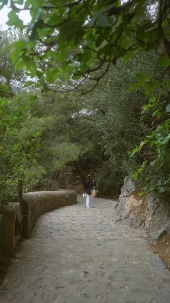 stock image Woman walking along a scenic forest path in mallorca, spain, surrounded by lush greenery and stone walls under a canopy of trees.